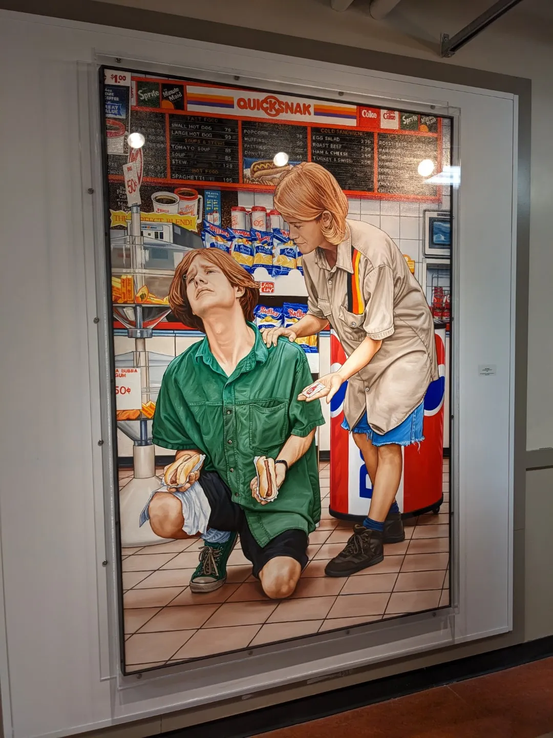 a painting by Chris Woods titled “The Realm of Convenience II”. It depicts a blonde man on his knees with hotdogs in his hands being consoled by another blonde woman with ketchup in her hand. They’re located in a convenience store.